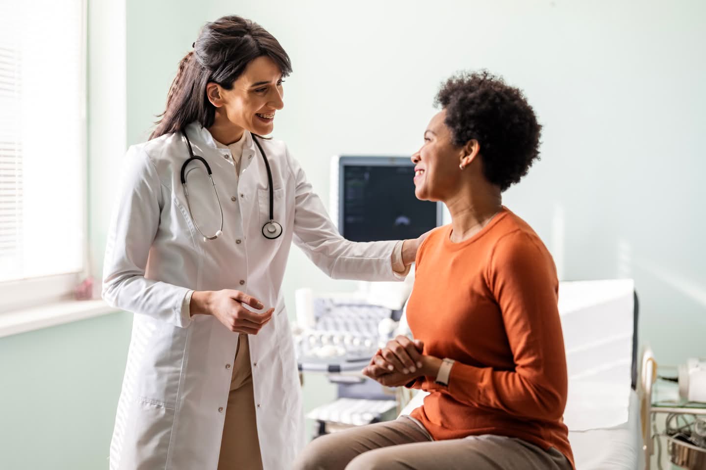 Female doctor smiling and patting the shoulder of a female patient seated on a bed, representing Cogstate