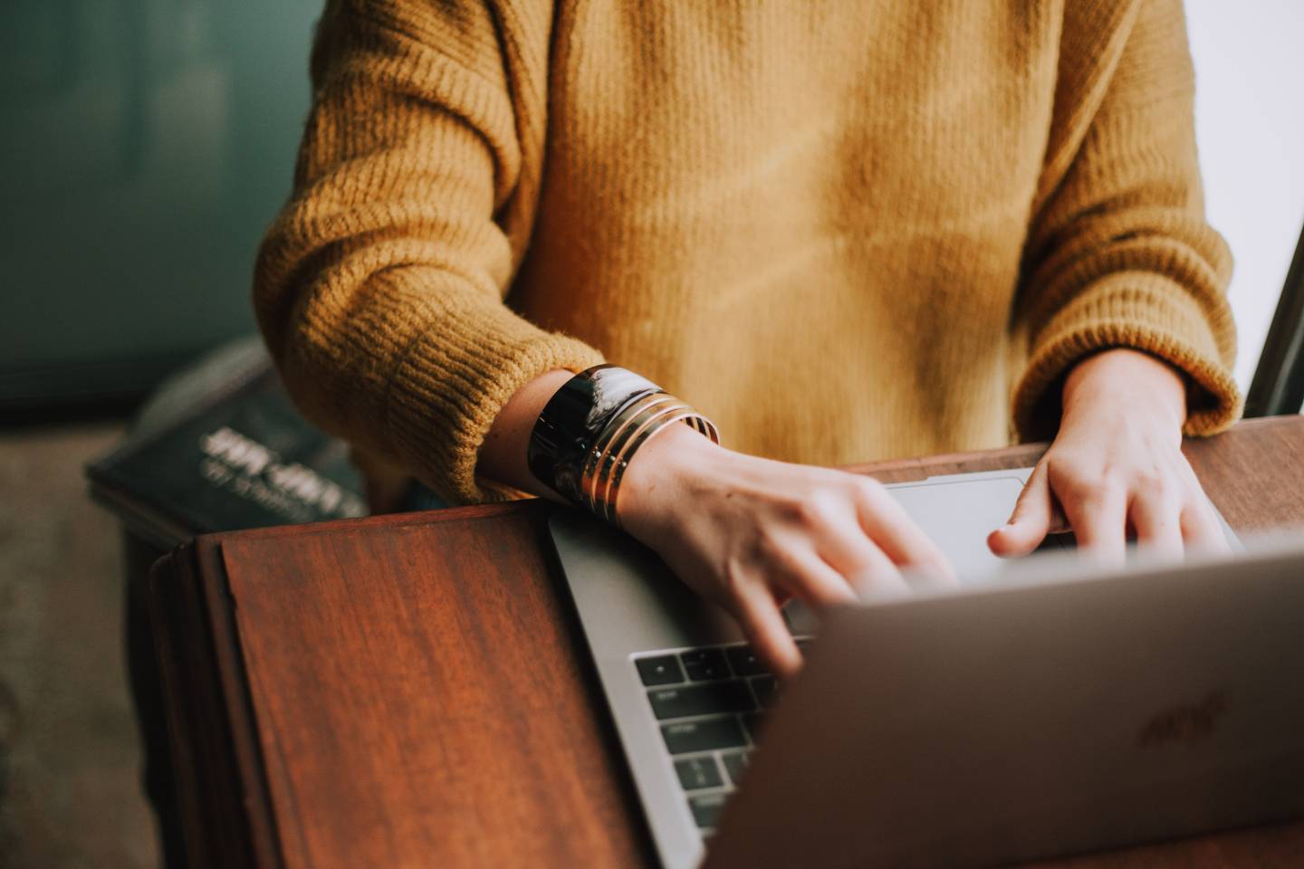 A person's hands typing on a laptop keyboard, wearing a chunky yellow sweater and a dark bracelet.