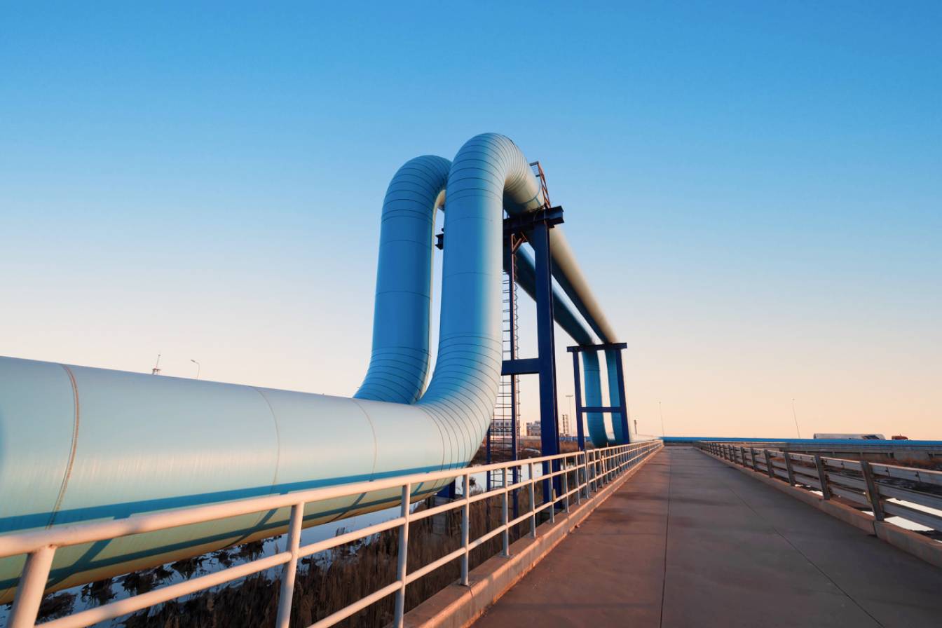 Large, light blue industrial pipeline curving steeply upward over a bridge or walkway, against a clear blue and orange sunset sky.