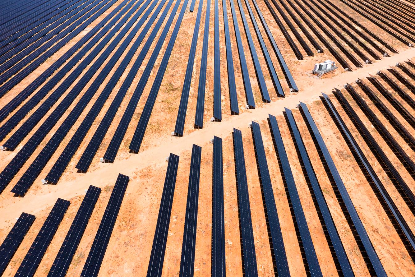 Aerial view of numerous rows of black solar panels arranged in parallel lines on dry, reddish-brown ground, with a small structure visible near the right side of the image.