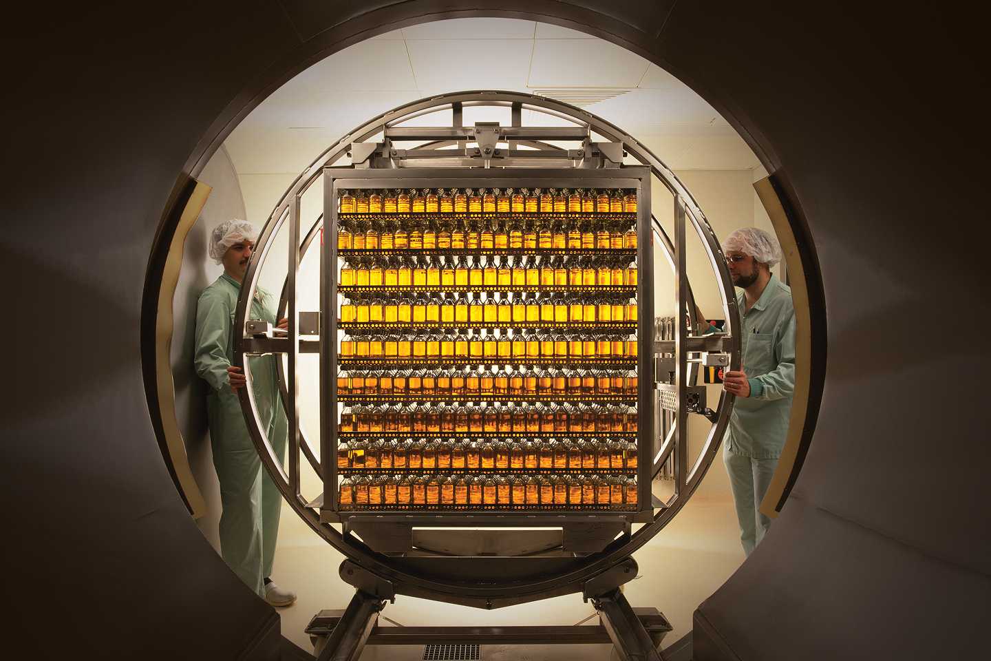 Two people in a lab wearing jumpsuits and hair nets bring bottles out of a vault